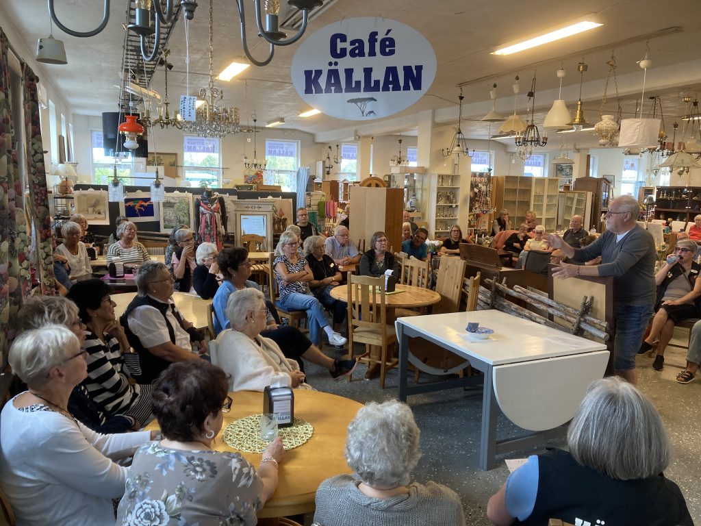 Besökare samlade i Café Källan medan en man håller ett anförande i secondhandbutiken